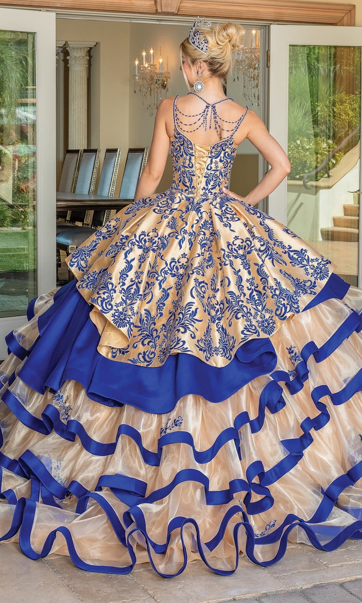 Dressed in the Quinceanera Dress 1689 By Dancing Queen—a gold and blue gown featuring intricate embroidery, a tiered ballgown skirt, and a sparkling tiara—this individual stands with their back to the camera before glass doors, embodying graceful elegance.
